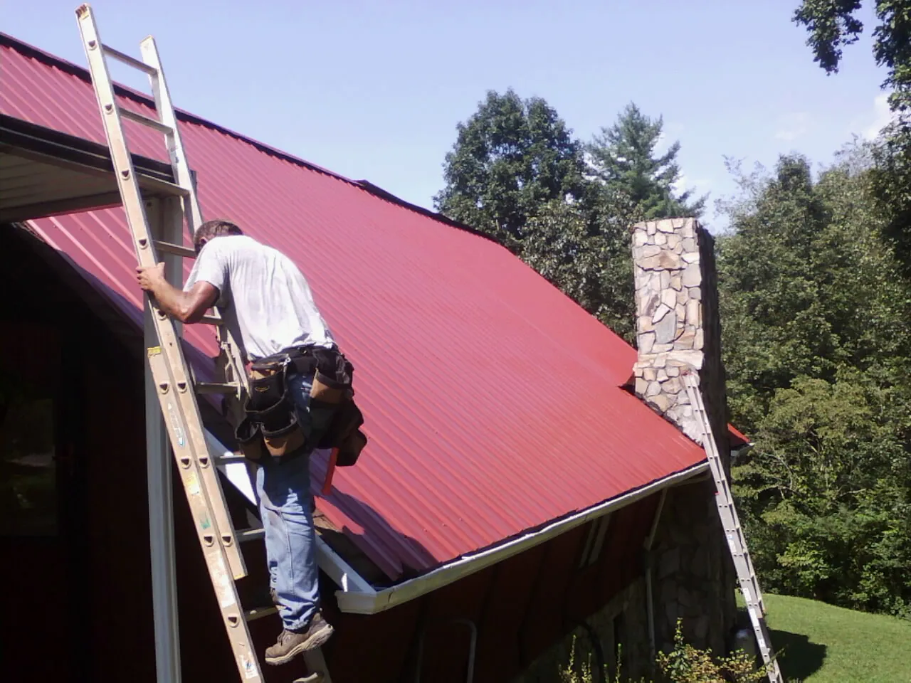 Roofer on a ladder beside a red metal roof and stone chimney — Soffit & Fascia Repair by Roofing Brothers in Whaleyville